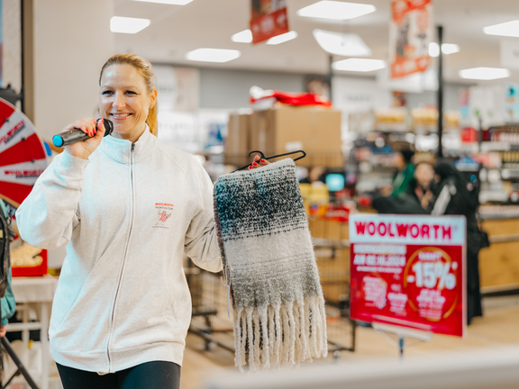 Eine lächelnde Frau hält ein Mikrofon und einen Schal in einem Geschäft mit Werbepostern im Hintergrund.