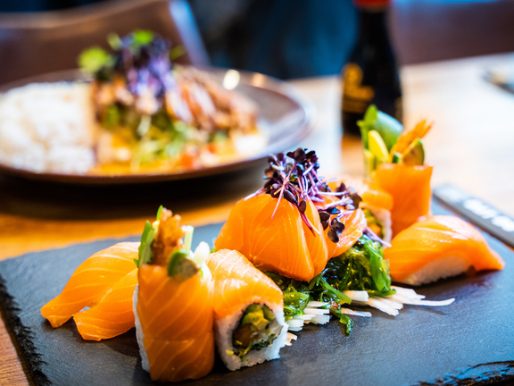 Sushi-Platte mit lila Kräutern und frischem Lachs auf schwarzem Teller, im Hintergrund eine weitere Speise.