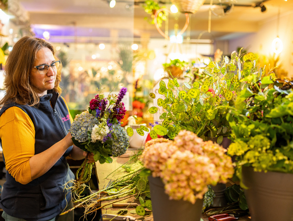 Frau in einem Blumenladen arrangiert bunte Blumen und Pflanzen vor einem Verkaufstresen. Warmes Licht im Hintergrund.