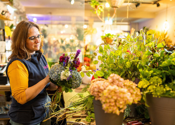 Frau in einem Blumenladen arrangiert bunte Blumen und Pflanzen vor einem Verkaufstresen. Warmes Licht im Hintergrund.