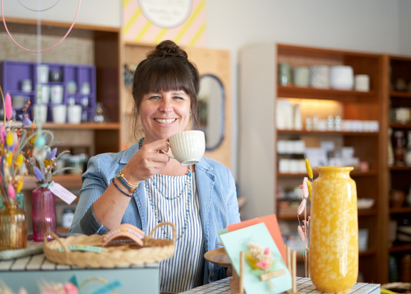 Frau mit Tasse in einem stilvollen Geschäft, umgeben von dekorativen Gegenständen und Pflanzen.