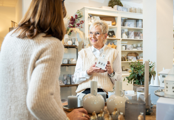 Eine lächelnde Verkäuferin zeigt einer Kundin ein Produkt in einem geschmackvoll eingerichteten Geschäft.