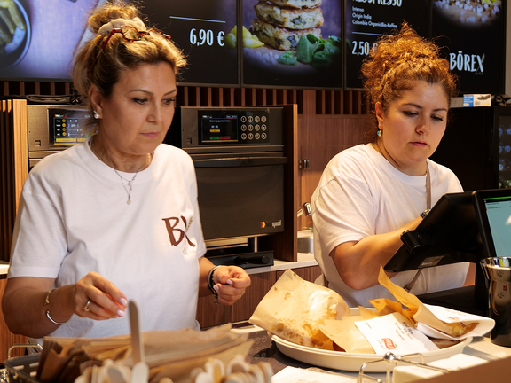 Zwei Mitarbeiterinnen in einem modernen Fast-Food-Restaurant bereiten Speisen vor und verwenden ein Tablet an der Theke.
