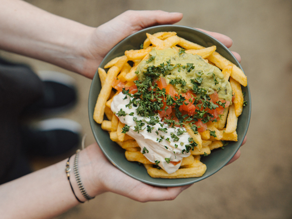 Eine Person hält eine Schüssel mit Pommes, Guacamole und einer cremigen Sauce, garniert mit frischen Kräutern.