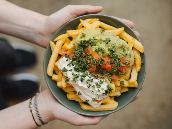 Eine Person hält eine Schüssel mit Pommes, Guacamole und einer cremigen Sauce, garniert mit frischen Kräutern.