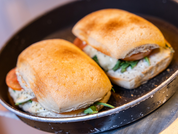 Zwei frisch zubereitete Sandwiches in einer Pfanne, auf einem Teller, mit gegrilltem Gemüse und frischen Kräutern.