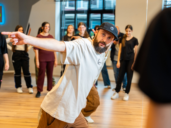 Tanztrainer zeigt während einer Klassenübung auf die Teilnehmer im Hintergrund, die in verschiedenen Outfits stehen.