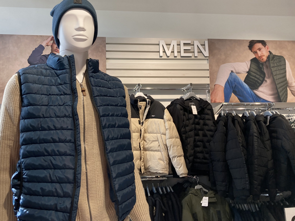 Mannequin in a dark blue vest and a beanie, displayed among men's outerwear, with a "MEN" sign above.