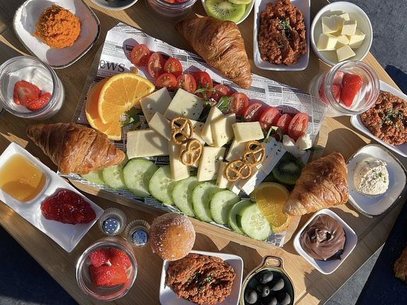Auf einem Holzbrett angeordnetes Frühstücksbuffet mit Croissants, Käse, Obst, Gemüse und Dips in Schalen.