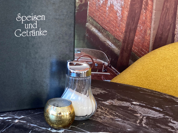 Schwarze Speisekarte auf einem marmorierten Tisch mit einem Salzstreuer und einem Kerzenhalter, im Hintergrund Wasser und Ziegel.