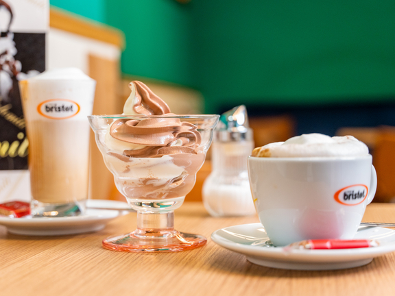 Eisbecher mit Schokoladeneis, Becher mit cremigem Getränk und einer Tasse Kaffee auf einem Tisch, im Hintergrund ein Menü.