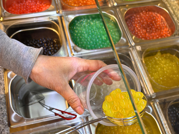 Eine Hand hält eine Schüssel mit gelben Perlen über mehreren Behältern mit bunten, runden Gel-Perlen.