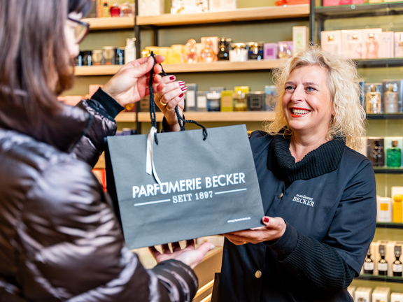 Frau überreicht eine Einkaufstüte in einem Parfümgeschäft an eine Kundin. Hintergrund mit Parfümflaschen.